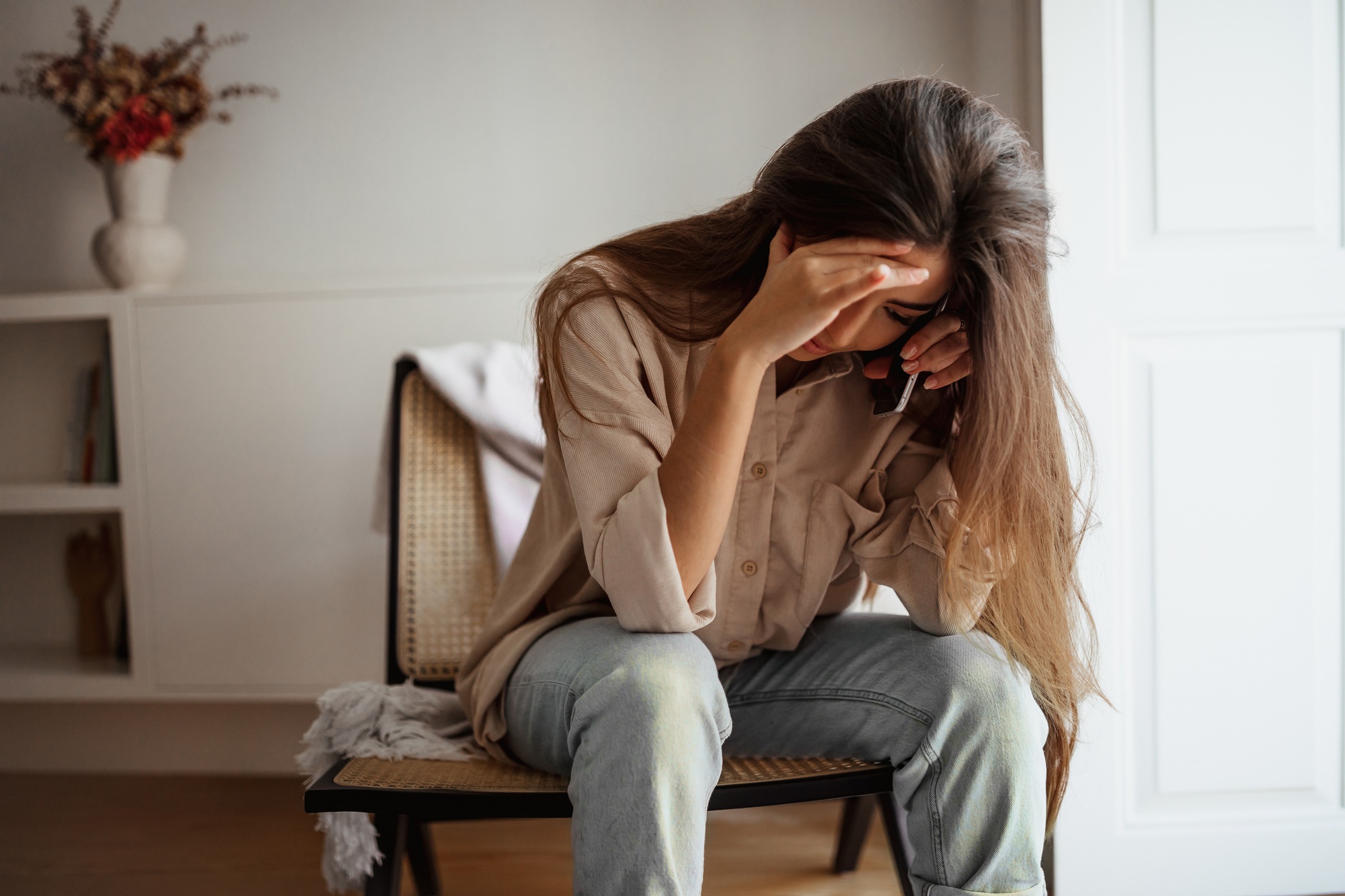 Despaired sad millennial mixed race female calls by phone, talks, sits on chair, suffering from bad news