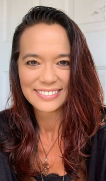 A woman with long, wavy reddish-brown hair smiles at the camera. She is wearing a black top and a necklace, and is in front of a light-colored background.