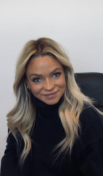 A woman with long, wavy blonde hair wearing a black turtleneck sweater sits on a dark chair, smiling at the camera against a plain white background.