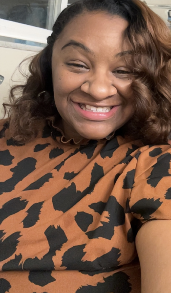 A woman with curled brown hair smiles brightly at the camera. She is wearing a brown and black animal print top, and the background includes a window and a cream-colored wall.